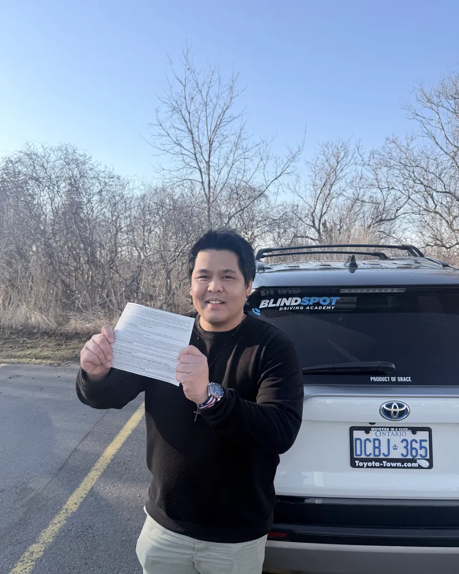 Student in a dark jacket beside the Blindspot Driving Academy car, holding their road test result