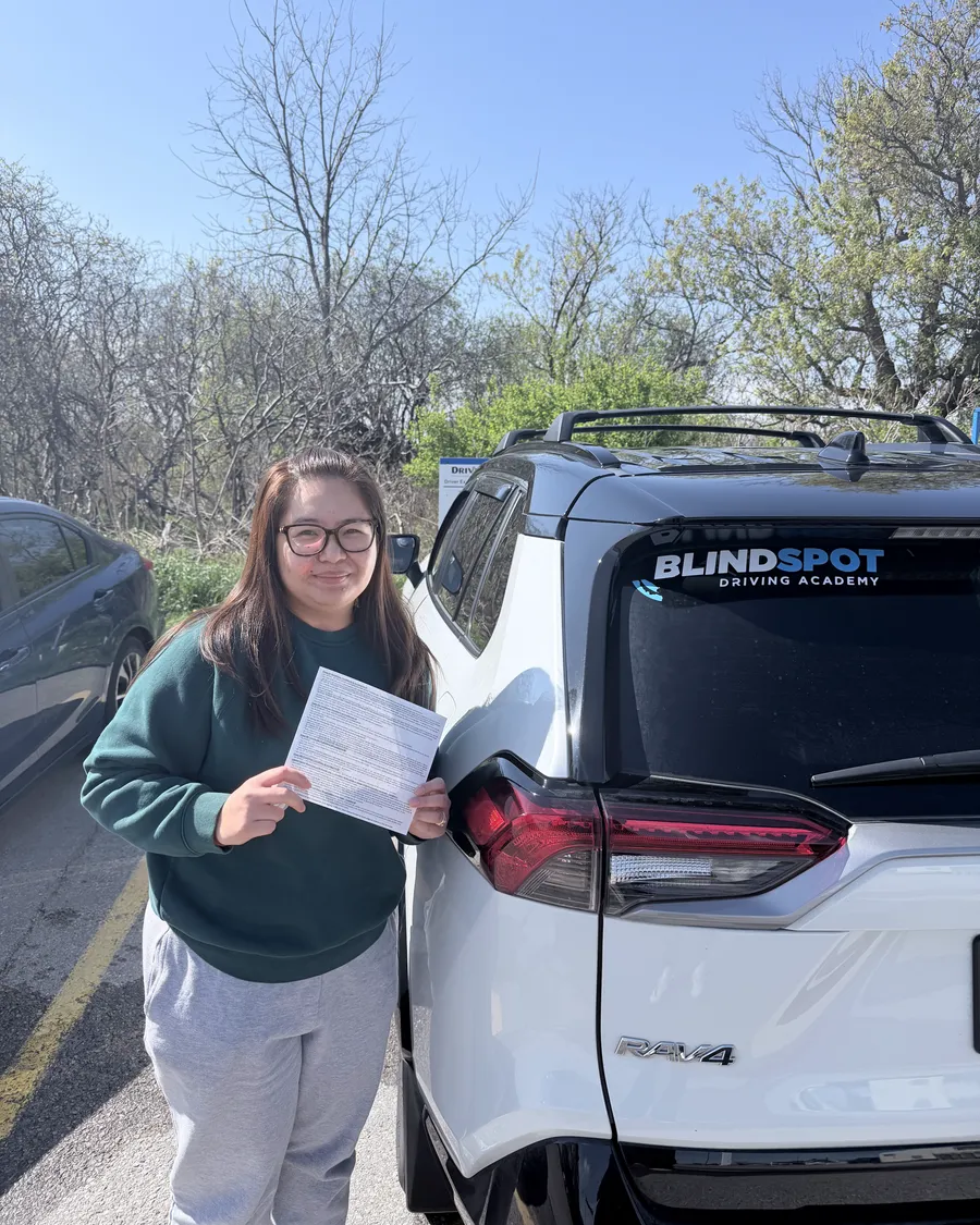 Student in a green sweater beside the Blindspot Driving Academy car, holding their road test result
