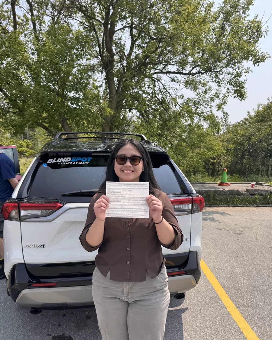 Student in a brown top beside the Blindspot Driving Academy car, holding their road test result