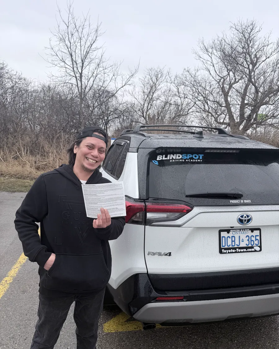 Student in a dark hoodie and beanie beside the Blindspot Driving Academy car, holding their road test result