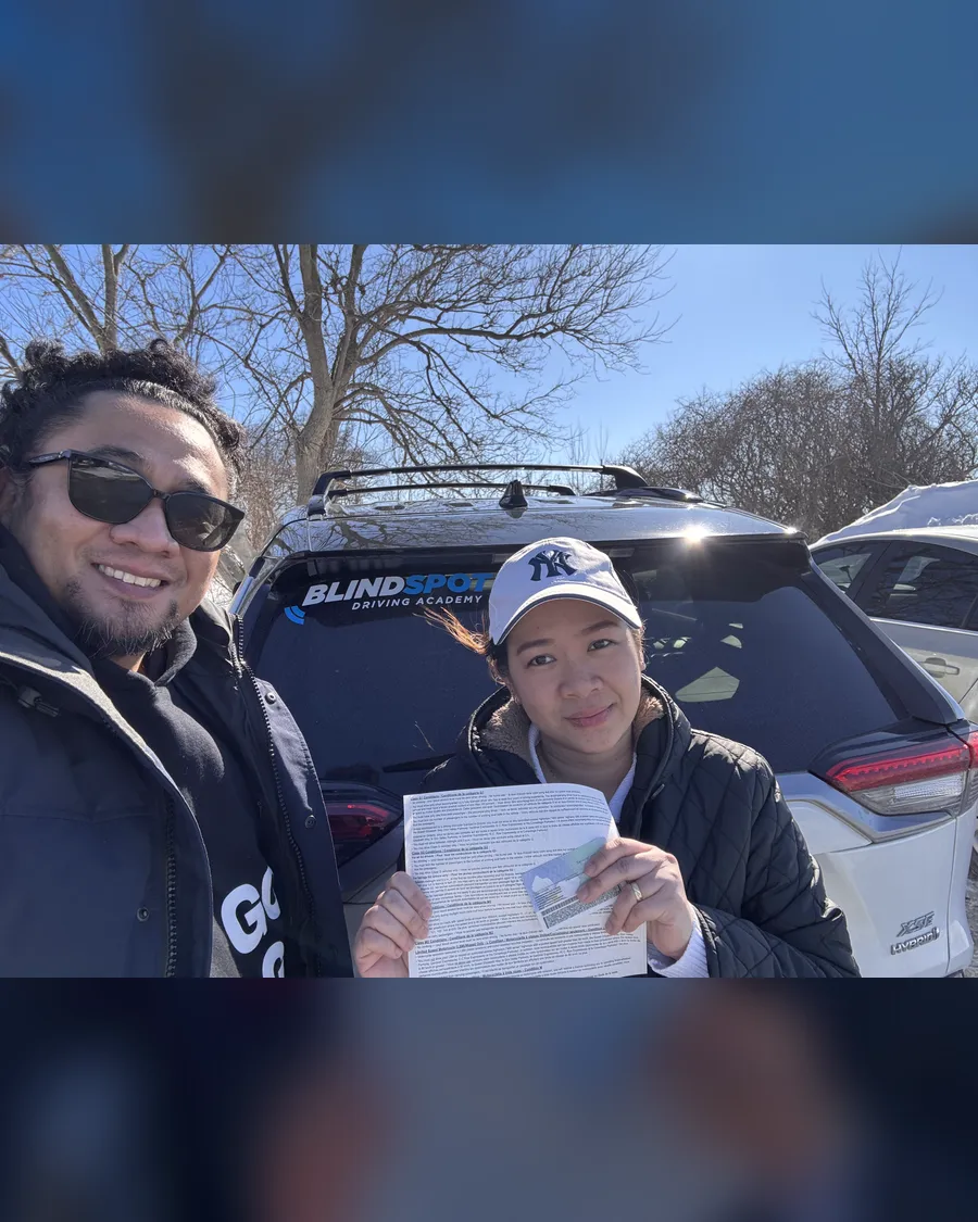 Student in a baseball cap and instructor Allan beside the Blindspot Driving Academy car, holding a road test result