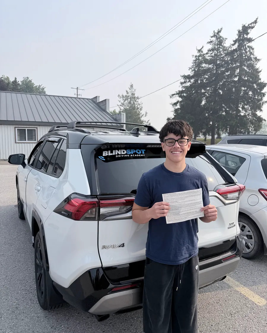Student in a navy t-shirt beside the Blindspot Driving Academy car, holding their road test result