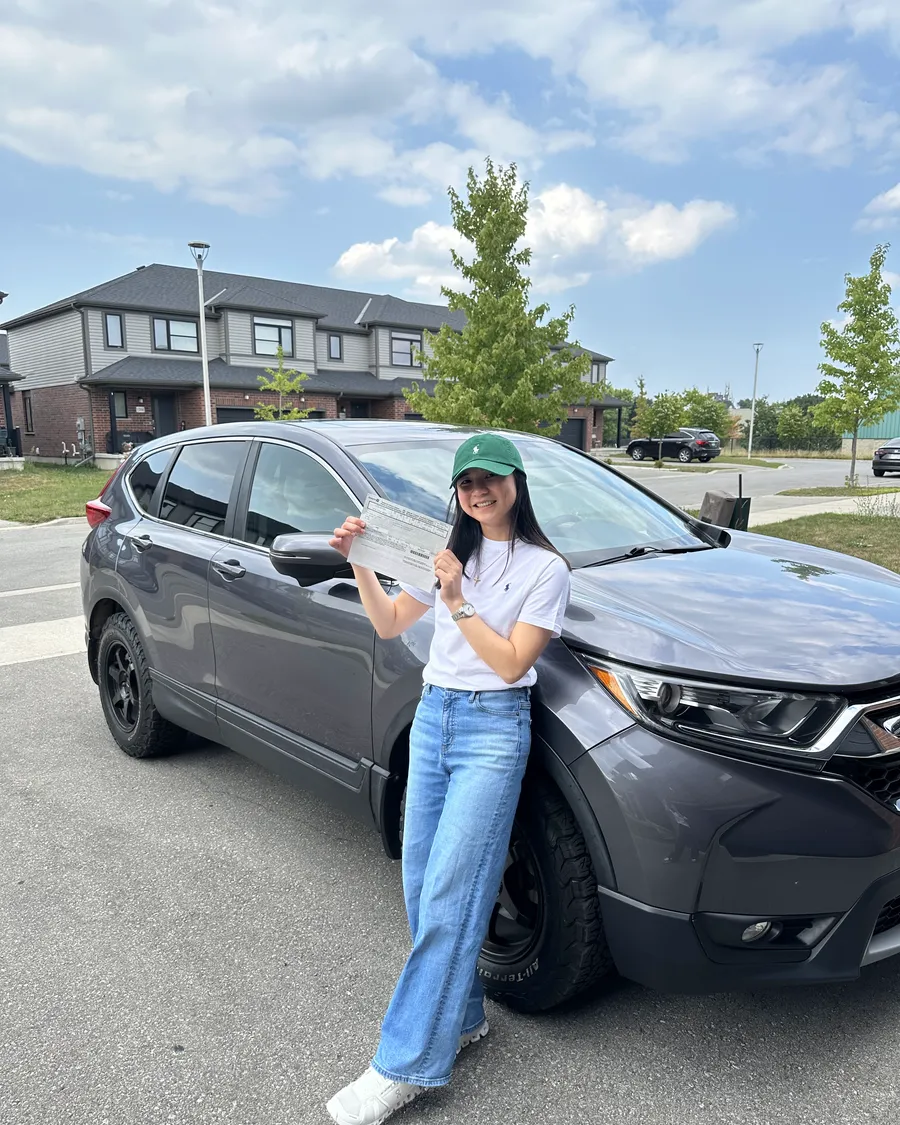 Student in a white top and green cap beside a car on a sunny day, holding their road test result