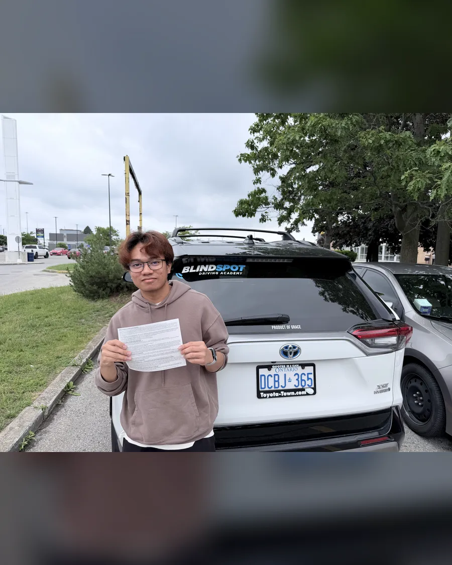 Student in a brown hoodie beside the Blindspot Driving Academy car, holding their road test result